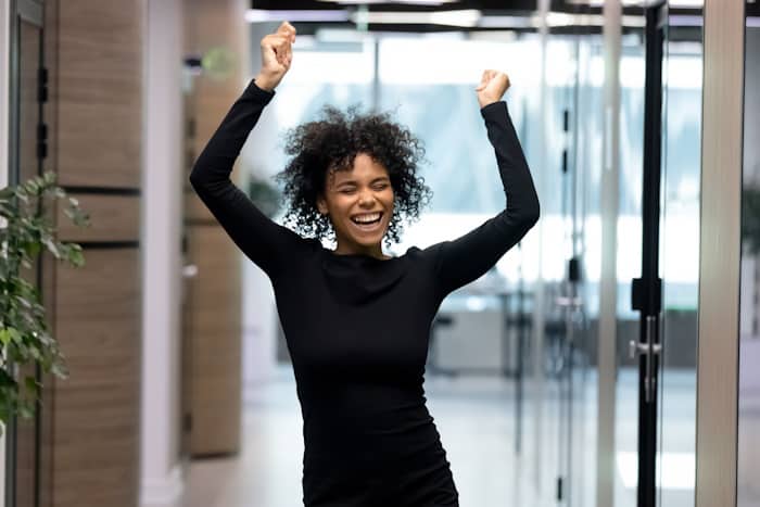 Overjoyed African American female employee dance celebrating success Overjoyed young African American female employee or worker dance in office hallway enjoy Friday fun. Happy smiling millennial biracial businesswoman celebrate job success or work achievement.