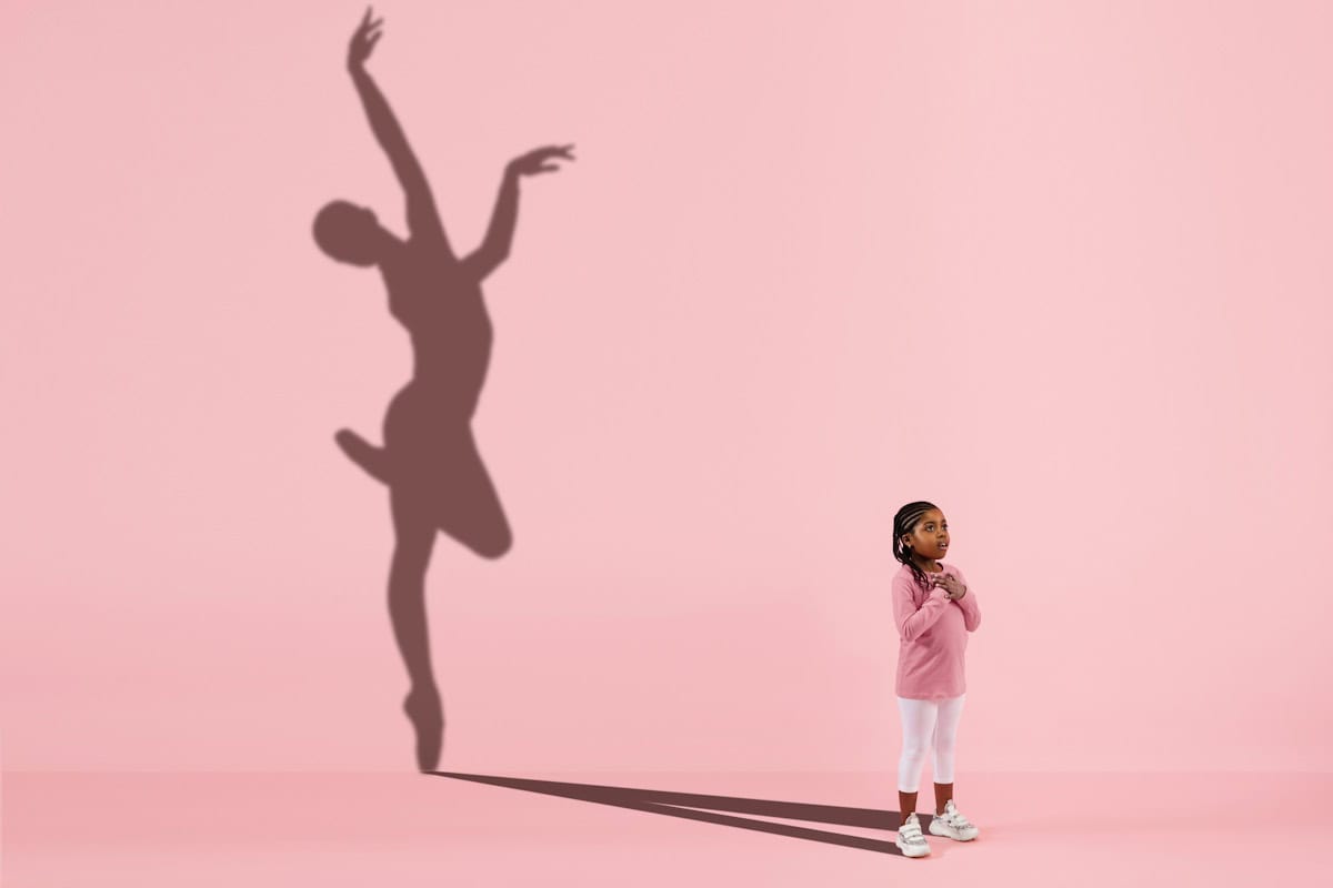Childhood and dream about big and famous future. Conceptual image with girl and drawned shadow of female ballet dancer on coral pink background. Childhood, dreams, imagination, education concept.